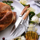 A roasted turkey on a white platter sits beside the Messermeister Oliva Elite Kullens Carving Set—2 Piece, featuring olive wood handles. White roses and a glass of amber-colored drink complete this elegant dining scene.