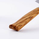 Close-up of the Messermeister Oliva Elite Kullens Santoku Knife - 7", featuring a smooth, polished olive wood handle with natural grain on a white background. The blade, showing its hollow edge, is partially visible in the top right corner.