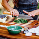 Wearing a gray apron, a person chops fresh herbs on a wooden board using the 7" Messermeister Oliva Elite Kullens Santoku Knife. A green bowl, a jar, and vegetables sit nearby on the table.