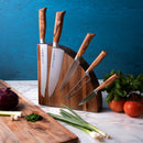 The Messermeister Oliva Elite Magnet Block Set - 6 Piece, featuring German forged knives with wooden handles, sits on a marble counter near chopped green onions, red onion, tomatoes, and basil with a blue background.