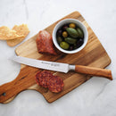 A wooden cutting board with sliced salami, a whole salami, the Messermeister Oliva Elite Reverse Scalloped Utility Knife - 6", mixed olives in a small bowl, and crisp bread pieces are arranged on a white surface.
