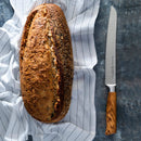 A loaf of seeded bread sits on a white striped cloth next to the Messermeister Oliva Elite Scalloped Bread Knife - 9", featuring an olive wood handle, all arranged on a textured gray surface.