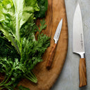 The Messermeister Oliva Elite Chef & Parer Set - 2 Piece features a wooden cutting board topped with leafy greens and two German high-carbon stainless steel knives, blades facing outward. Brand: Messermeister.