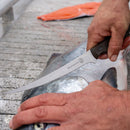 A person uses a fillet knife to cut a large fish on a wooden surface, one hand gripping the knife while the other steadies the fish. Nearby, the Messermeister Overland Chef 6-Piece Leather Knife Roll Set from Messermeister securely holds an assortment of outdoor cooking knives, ready for any culinary adventure.