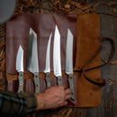 A hand selects a knife from the Messermeister Overland Chef 6-Piece Leather Knife Roll Set—a premium leather roll by Messermeister holding five different outdoor cooking knives—laid out on a wooden surface with dried leaves scattered around.
