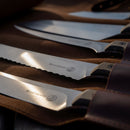 Six Messermeister knives, each engraved with the brand’s signature on the blades, are neatly arranged in the Messermeister Overland Chef 6-Piece Leather Knife Roll—their polished metal catching the sunlight—making this set an ideal companion for any outdoor cooking adventure.