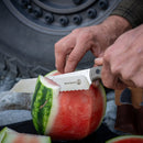 Outdoors, a person slices a watermelon using a serrated knife from the Messermeister Overland Chef 6-Piece Leather Knife Roll Set. The Messermeister brand is visible on the blade, indicating it is part of their premium outdoor culinary collection. In the background, there’s a large tire and rocky ground.