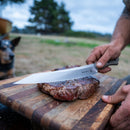 Outdoors, a person slices a cooked steak on a wooden cutting board with a large knife from their Messermeister Overland Chef 6-Piece Leather Knife Roll Set; the scene includes a dog and trees softly blurred in the background.