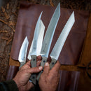 A person holds three large, shiny Messermeister knives with textured handles, while a fourth knife and the brown leather Messermeister Overland Chef 6-Piece Leather Knife Roll are visible in the background on a wooden surface.