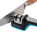 A kitchen knife with a wooden handle is being sharpened using the Messermeister Pull Through Sharpener by Messermeister, which features a black and blue base.