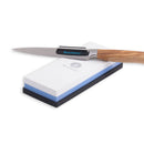 A paring knife with a wooden handle rests on the Messermeister Polishing Stone—2000 & 5000 grit—with a white top layer, blue base, and Messermeister logo, perfect for precise sharpening and polishing.