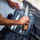 A person selects a chef’s knife with a wooden handle from the Messermeister Preservation Heather Gray Knife Roll (5 Pocket) on a rustic wooden table, with kitchen essentials and a cloth nearby.