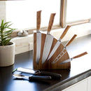 A Messermeister Wood Quarter Round Magnetic Knife Block by Messermeister holds five wooden-handle knives on a black countertop near a window and potted plant, with three black-handled utensils in front—an elegant solution for stylish kitchen knife storage.