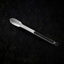 A Messermeister Stainless Locking Tong, 12", with scalloped edges and a black handle grip rests on a dark, textured surface.