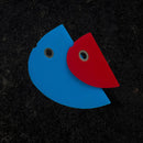 Two flat, semi-circular silicone shapes—one blue and one red, each with a hole near the edge—overlap on a dark surface. These are the Messermeister 2 Piece Silicone Bowl Scraper Set, essential kitchen utensils from Messermeister.