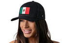 A woman with long brown hair smiles, wearing a Haka Hat Mexican Flag Trucker Hat (Black Mexico Snapback) by Haka Hat. The background is white.