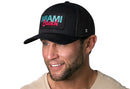 A man wearing the Haka Hat Miami Beach Trucker Hat in black, featuring a teal and pink patch. He smiles, showing blue eyes, while the snapback from Haka Hat gives his look a relaxed vibe.