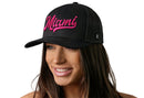 A woman with long brown hair smiles while wearing the Haka Hat Miami Hot Pink Script Trucker Hat, a black Florida snapback by Haka Hat featuring "Miami" in hot pink script on the front. The background is white.
