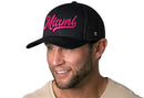 A man with a trimmed beard wears the Haka Hat Miami Hot Pink Script Trucker Hat—black Florida snapback—paired with a light beige shirt, against a white background.