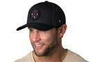 A smiling man with light stubble wears the Haka Hat Miami Trucker Hat | Black MIA X Snapback, featuring a circular MIA patch and blue gun icon. He pairs it with a beige shirt against a white background for a classic, stylish look.
