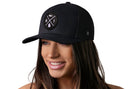 A woman with long brown hair is smiling and wearing the Haka Hat Michigan Trucker Hat | Black MI X Snapback, featuring a round patch with Michigan symbols. The background is white.