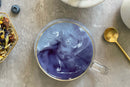 A glass cup filled with swirling purple and white World Spice Midnight Berry Tea from World Spice sits on a gray surface, next to a gold spoon, a bowl, and a dish of dried tea leaves and blueberries.