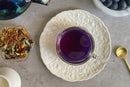 A clear teacup filled with World Spice Midnight Berry Tea by World Spice sits on a white floral plate, surrounded by dried tea leaves, a bowl of blueberries, a golden spoon, and part of a teapot on a gray surface.