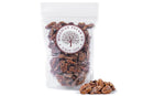 A clear, resealable 12 oz bag of Millican Pecan Cinnamon Candied Pecans features the Millican Pecan brand label. In front of the bag, a few cinnamon-sugar coated Texas pecans are scattered, showcasing this irresistible snack.