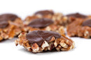 A close-up of Millican Pecan Dark Chocolate Caramillicans reveals luscious gourmet candies featuring a decadent caramel and pecan base, finished with a smooth layer of rich dark chocolate. Other Millican Pecan treats are softly blurred in the background atop a white surface, creating an elegant presentation ideal for any gourmet chocolate gift box.