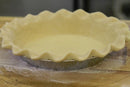 A Millican Pecan Chocolate Pecan Pie—9 Inch Southern Dessert from Millican Pecan, featuring a flaky unbaked crust with fluted edges in a metal pie tin, sits on a floured wooden surface, ready for filling and baking—the ideal beginning for your classic Southern chocolate pecan pie.