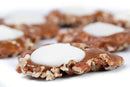 Close-up of Millican Pecan White Chocolate Caramillicans – Individual from Millican Pecan, featuring cookies coated in caramel and white chocolate, reminiscent of classic turtle-style candy, with additional Caramillicans softly blurred in the background.