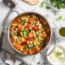 A pot of vegetable minestrone soup sits on a linen cloth, surrounded by SALTOPIA Infused Sea Salts' Tomato & Italian Herbs Seasoning: MYSTICAL PIZZA, grated cheese, basil leaves, olive oil, spoons, and slices of bread.