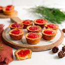 Nine small tartlets with golden crusts and red jam filling, made with Terrapin Ridge Farms Red Pepper Jelly (5.3oz), are arranged on a round wooden board, perfect as a cheese board condiment. One is bitten to reveal a creamy inside; pinecones and greenery accent the scene.