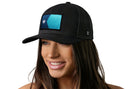 A woman with long brown hair smiles while wearing a black Haka Hat Minnesota Flag Trucker Hat featuring a blue patch and white star on the front. She stands against a plain white background.
