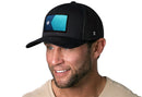 A man with a trimmed beard smiles, wearing the Haka Hat Minnesota Flag Trucker Hat— a black snapback with a blue geometric patch and white star— paired with a light beige shirt, set against a plain white background.