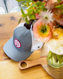 The Haka Hat Mom Trucker Hat, a gray-white snapback by Haka Hat with a red MOM patch, rests on a wooden table beside a vase of colorful flowers.