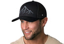 A man with a trimmed beard smiles in a beige shirt, wearing the Haka Hat Mountain Range Trucker Hat—an outdoors black snapback by Haka Hat with a mountain design on the front—against a plain white background.