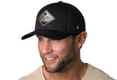 A smiling man wears the Haka Hat Diamond Mountain and Trees Trucker Hat in black by Haka Hat with a beige shirt, shown against a white background.