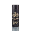 A black tube of The Immaculate Beard Mustache Wax Sweet Orange, made with natural ingredients and weighing .15oz (4.25g), Batch 1991, stands upright on a white background with a faint reflection below it.