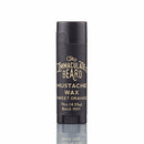A black tube of The Immaculate Beard Mustache Wax in Sweet Orange scent (.15oz), featuring vintage-style white lettering, stands upright against a white background—perfect for pairing with The Immaculate Beard Dark Shave Soap Puck to complete your grooming routine.