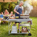 A man cooks at the Movable Grill Table with 2 Spice Containers and Station for Backyard and BBQ by DirectDeals, while his family and dog enjoy food and drinks at a nearby picnic table on a sunny day.