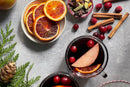 Two mugs of mulled wine with orange slices and cranberries, surrounded by cinnamon sticks, World Spice Mulling Spice from World Spice, additional cranberries, orange slices on a plate, greenery, and a pine cone on a gray background.