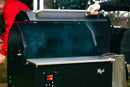 A person lifts the lid of a Myron Mixon Smokers BARQ-H2O Pellet Smoker, revealing smoke inside. The smoker uses H2O cooking technology and features a digital temperature display for precise competition-style BBQ.