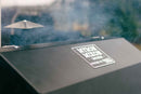 Close-up of a Myron Mixon BARQ-H2O Pellet Smoker outdoors, with smoke rising from the vent and the Myron Mixon Smokers logo visible on the front, highlighting competition BBQ and advanced H2O cooking technology.