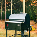 The Myron Mixon BARQ-H2O Pellet Smoker by Myron Mixon Smokers, featuring a digital control panel and side metal bucket, stands on grass with autumn leaves—perfect for competition-style BBQ.