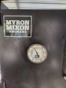 A Myron Mixon Smokers BBQ displays the 5" High Visibility Temperature Gauge by Myron Mixon Smokers, showing a 100°F–500°F range. The sleek black smoker sits partially under a metal roof, highlighting precise temperature control for ideal BBQ.