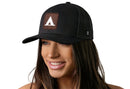 A smiling woman with long brown hair models the Haka Hat Tent Trucker Hat—a black mesh snapback by Haka Hat featuring a brown patch with a white triangle, inspired by campground trail signs—against a plain white background.