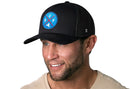 A man with a short beard wears a beige shirt and the Haka Hat Buffalo Trucker Hat | Blue Red BUF X | Black Snapback by Haka Hat, smiling as he looks to the side against a white background.