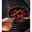 A hand holds the Napoleon Grills 9.5" Cast Iron Frying Pan (56053) with barbecue-glazed pineapple chunks above a hot grill, showcasing its grill-ready design and superior heat retention for outdoor cooking.