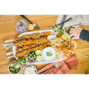 A platter of grilled chicken skewers, made using the Napoleon Rotisserie Shish-Kebab Skewer Set (64008) by Napoleon Grills, is served with naan, rice, cucumber, onion, jalapeños, lime, and sauces. A hand squeezes lime over the meal on a wooden table.
